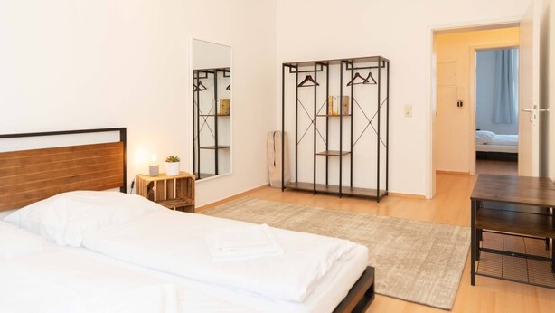 A bedroom with a bed, bedside table, mirror, wardrobe, and a view into another room.