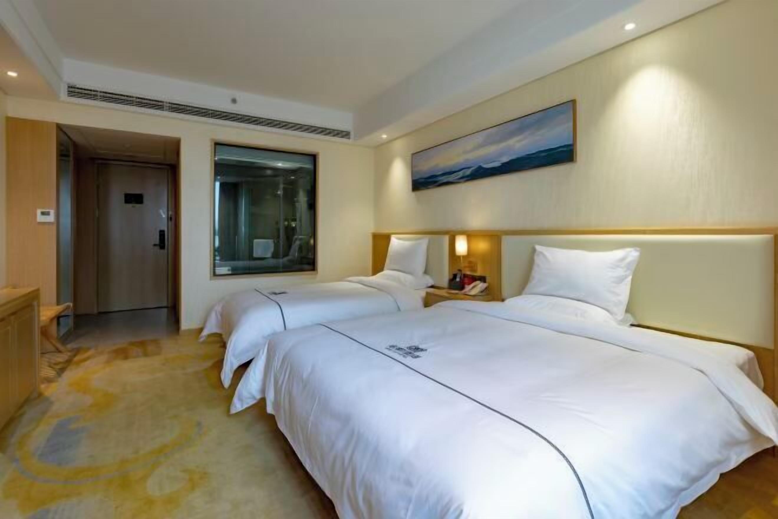 A hotel room with two beds, a large window, a painting on the wall, and a small table with a lamp.