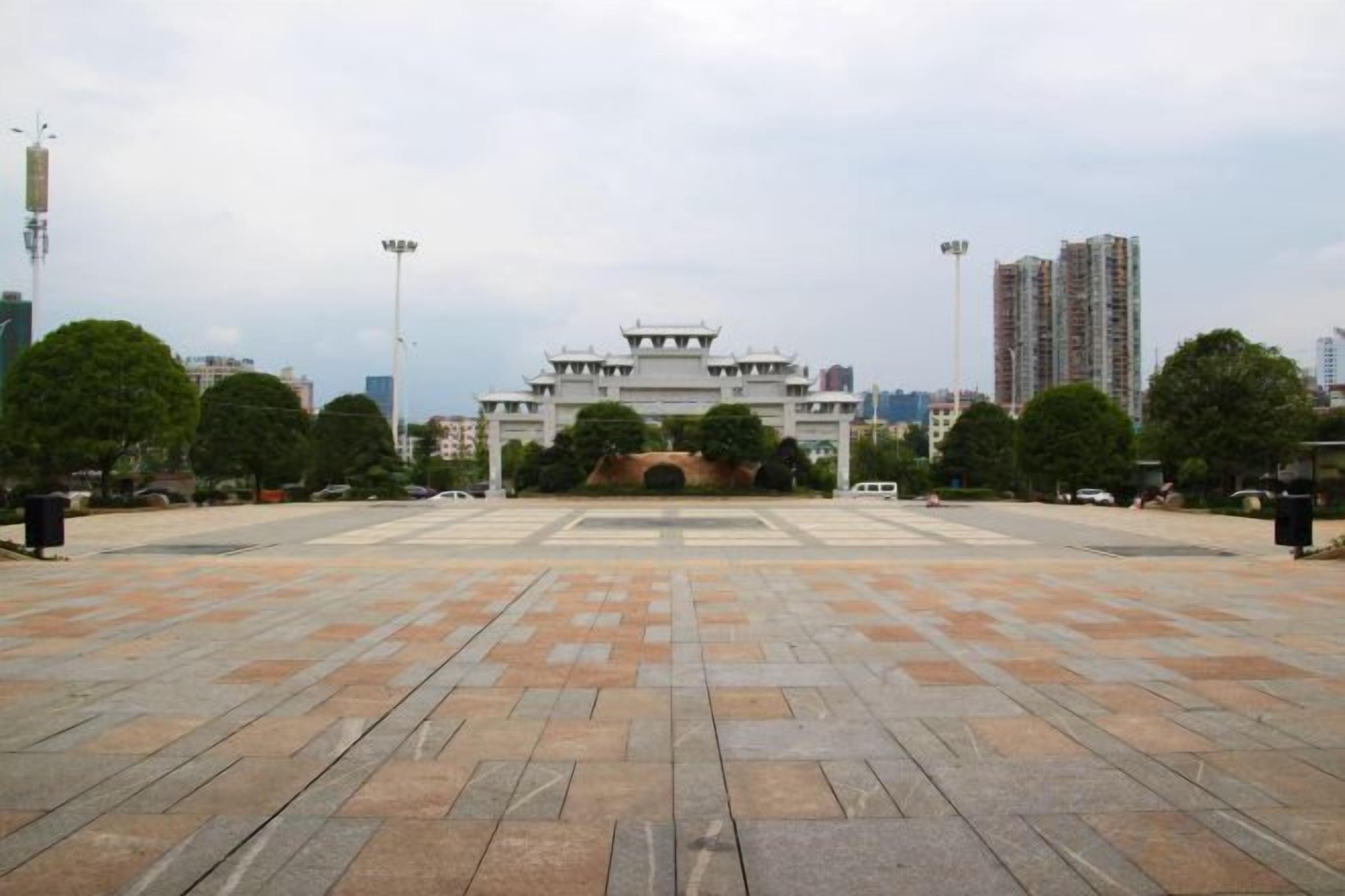 Una amplia plaza con un edificio tradicional al fondo, rodeada de árboles y modernos rascacielos.