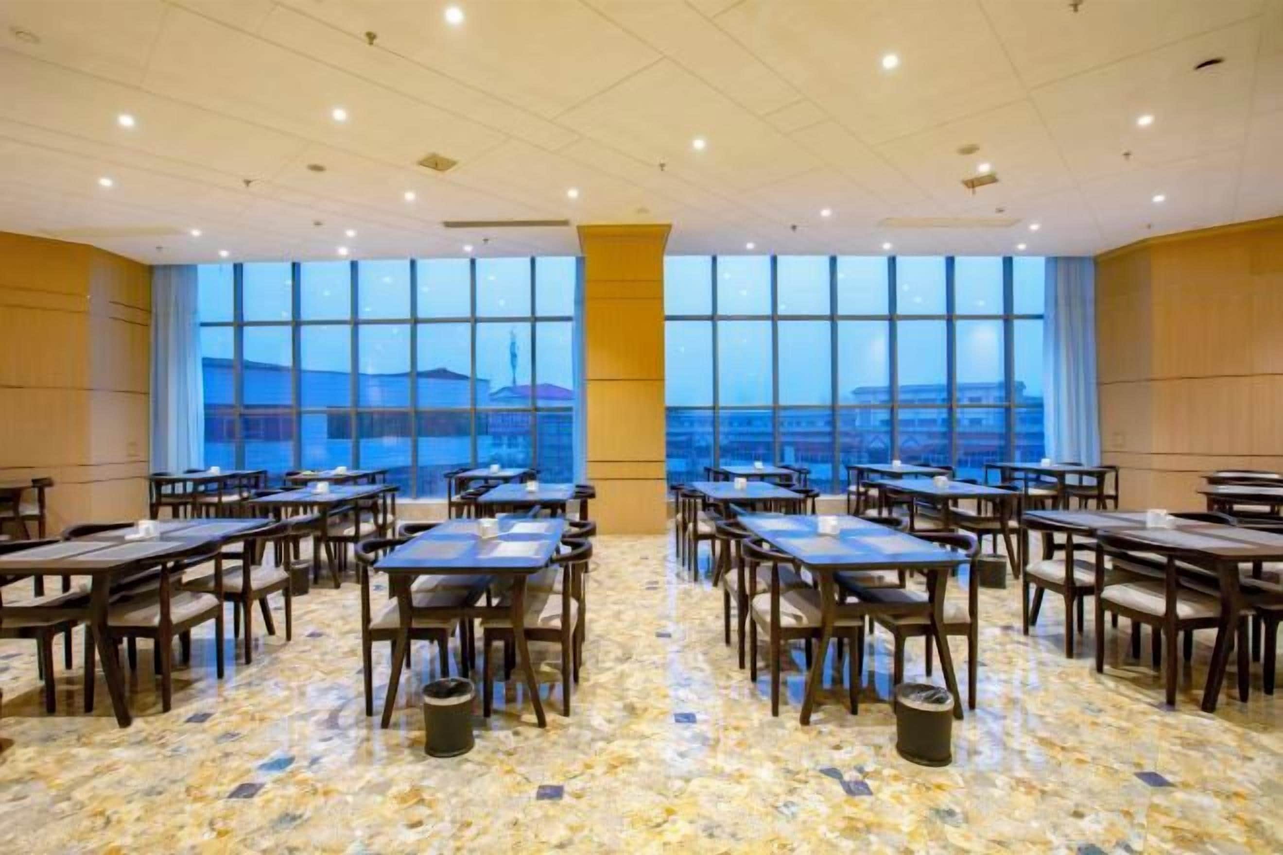 Un salón de conferencias con mesas y sillas dispuestas para reuniones, ventanas amplias con vistas y suelo de baldosas.