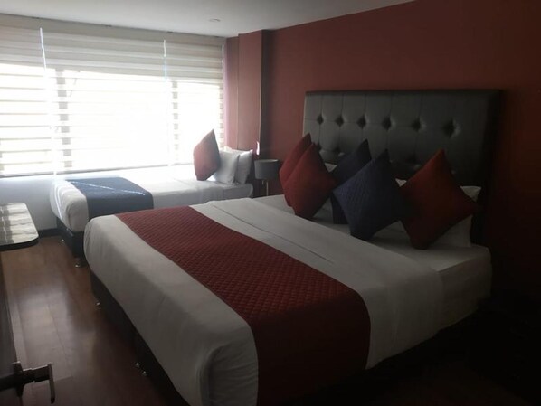 Een hotelkamer met twee bedden, een groot raam met jaloezieën en een rode accentmuur.