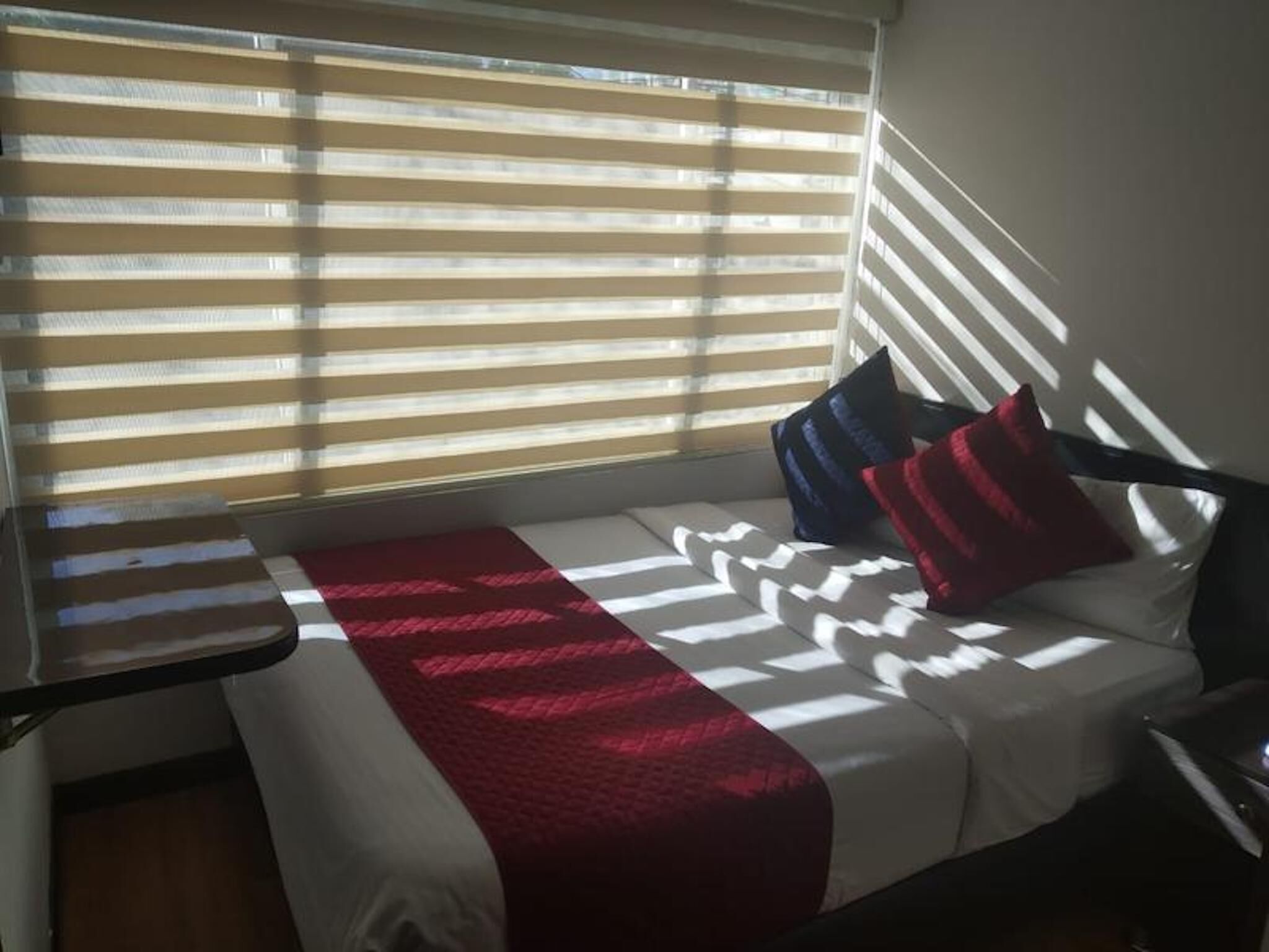 Una cama pequeña con una colcha roja y blanca, un almohadón y una mesita de noche.