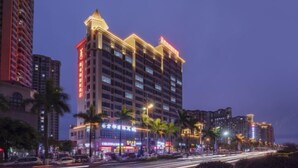 A multi-story hotel with a prominent corner entrance, illuminated signage, and a row of palm trees in front.