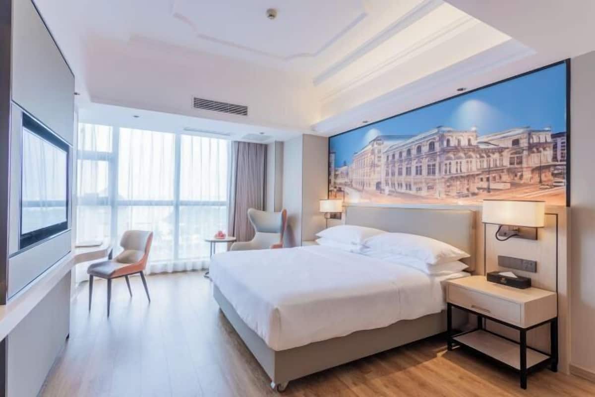 A hotel room with a large bed, a desk, a chair, a window, and a painting on the wall.