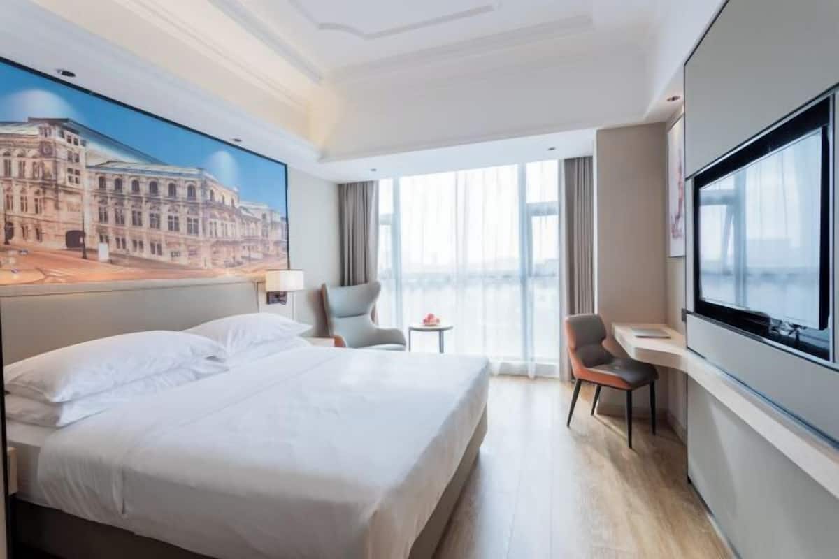A hotel room with a large bed, a desk, a chair, a TV, and a painting of a building on the wall.