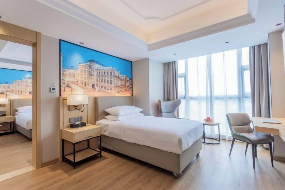 A hotel room with a bed, bedside tables, a desk, a chair, a window with curtains, and a large painting on the wall.