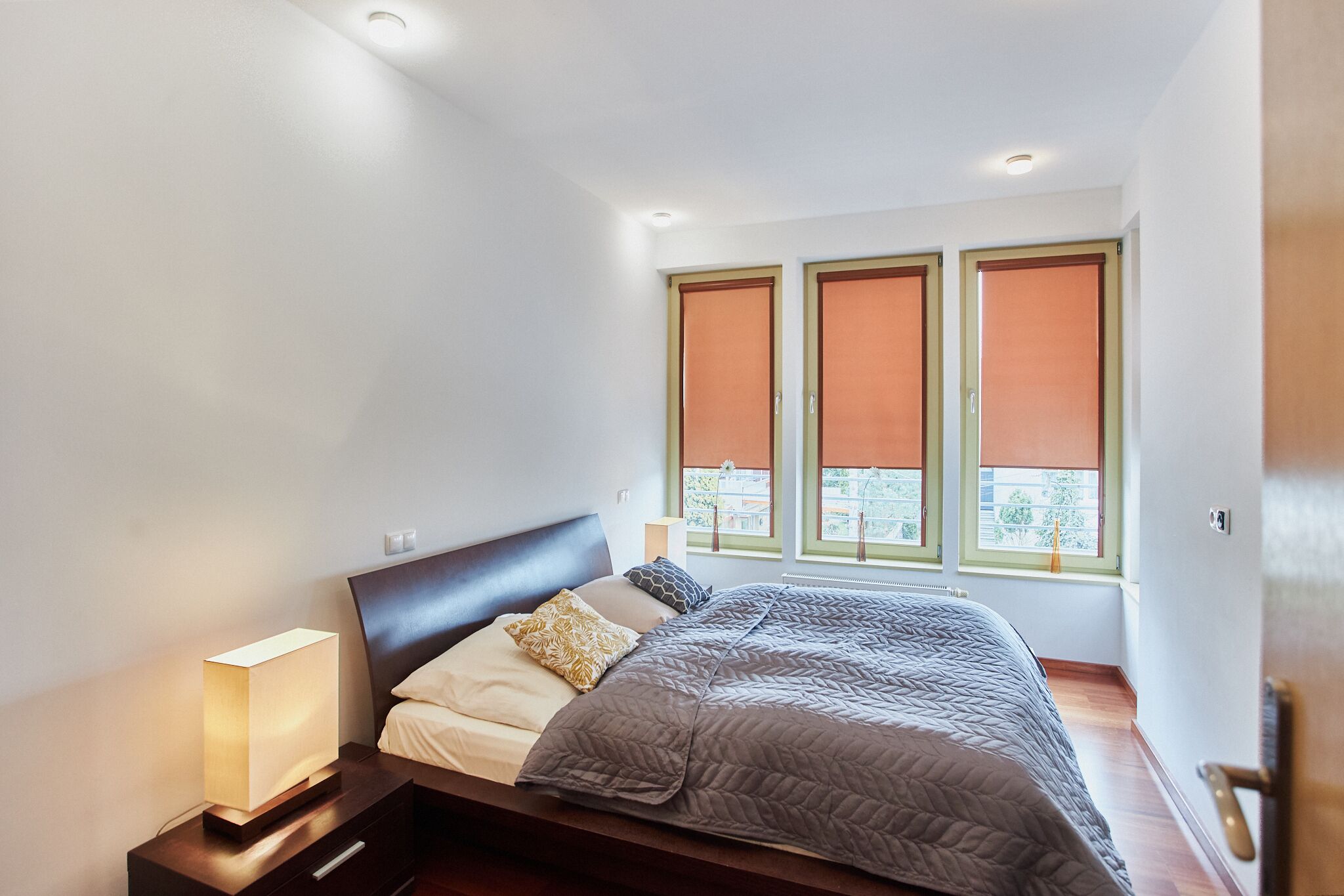 A modern bedroom with a bed, bedside lamp, and three windows with blinds.