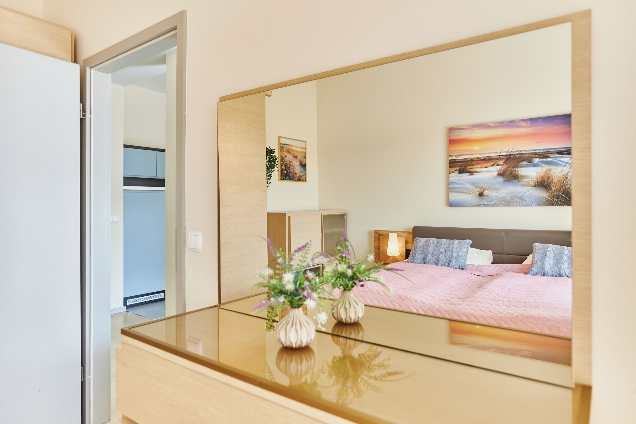 Une chambre avec un grand miroir, un lit recouvert d’une couverture rose et un tableau représentant un coucher de soleil accroché au mur.