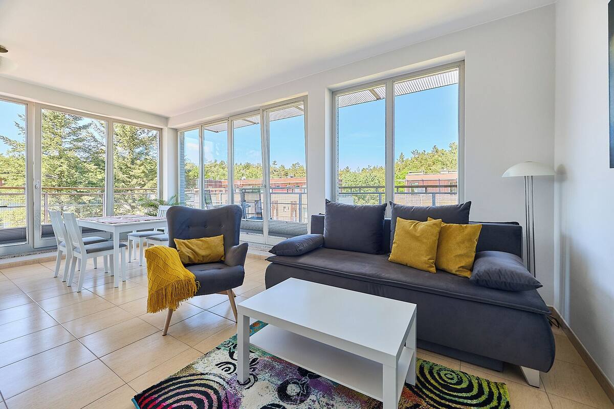 A modern living room with a grey sofa, two armchairs, a white coffee table, and a dining table with chairs. Large windows provide a view of the outdoors.