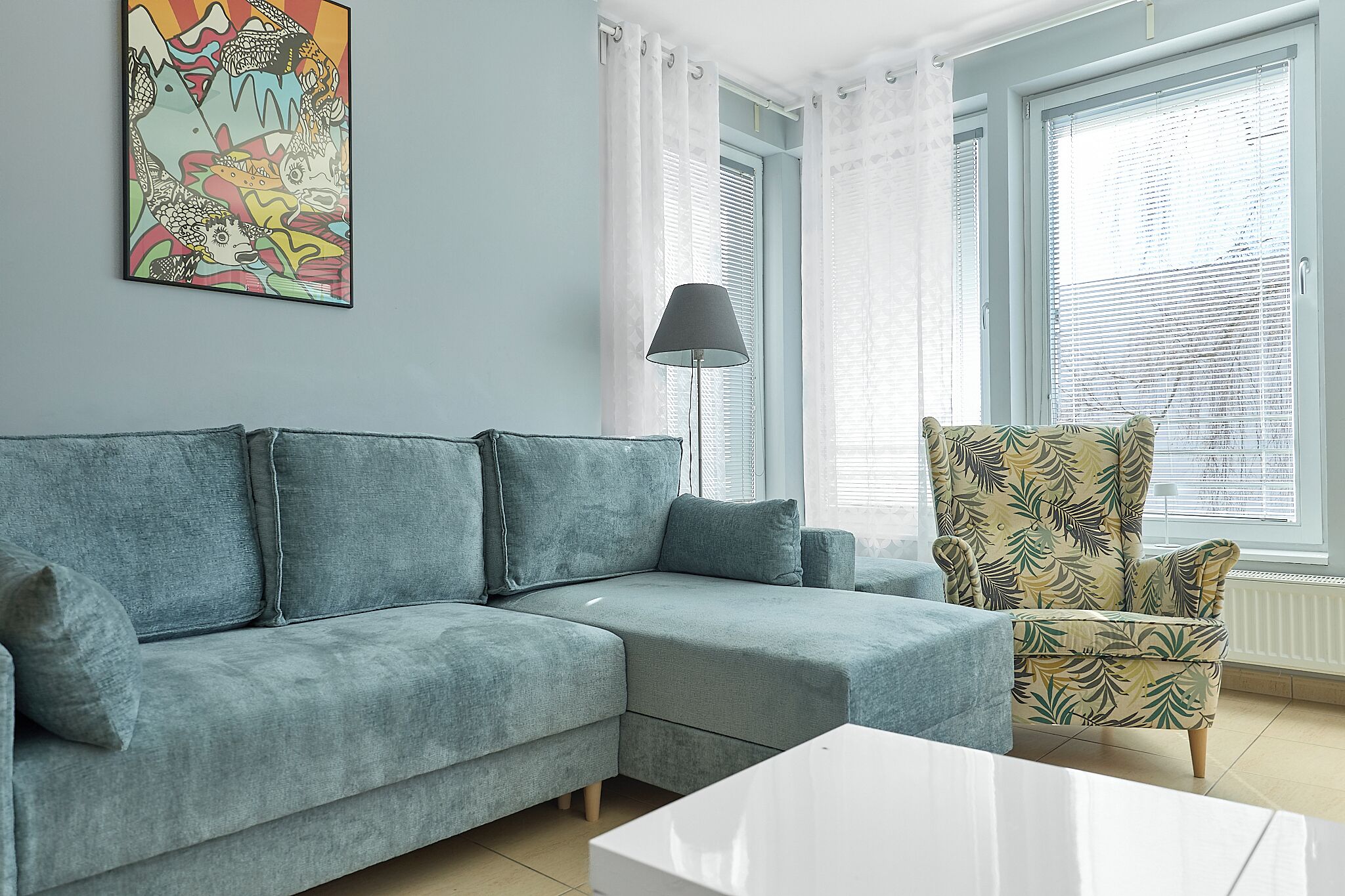 A living room with a grey sectional sofa, a patterned armchair, a white coffee table, and a colorful abstract painting on the wall.