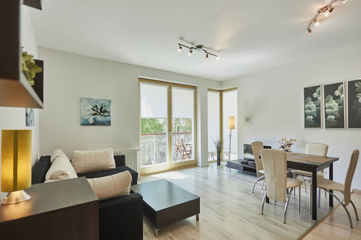 A modern living room with a sofa, coffee table, and dining table with chairs. There are large windows and a balcony visible.