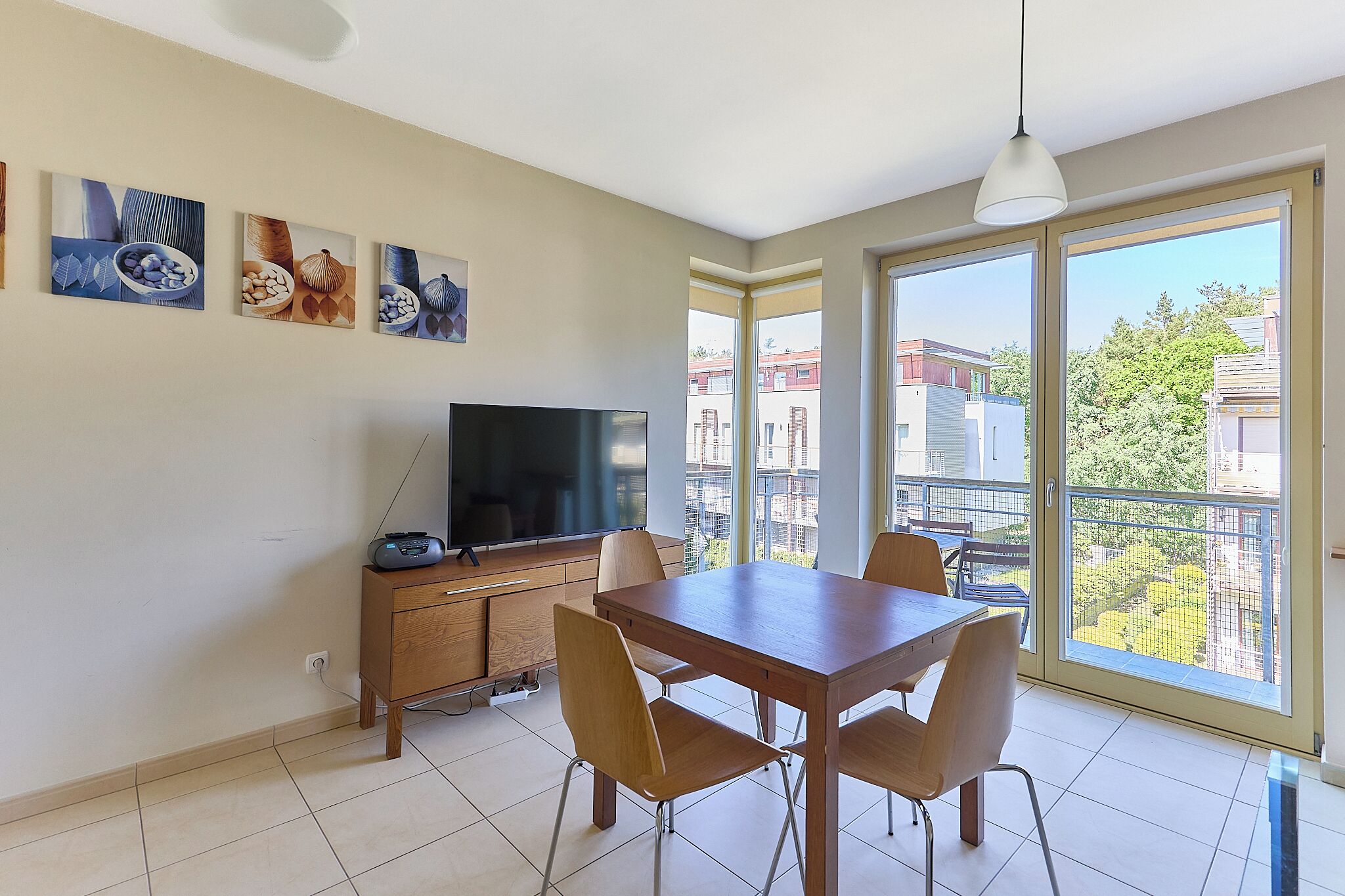 Un comedor con una mesa y sillas, un televisor sobre un mueble de madera y ventanas amplias con vistas a edificios y árboles.