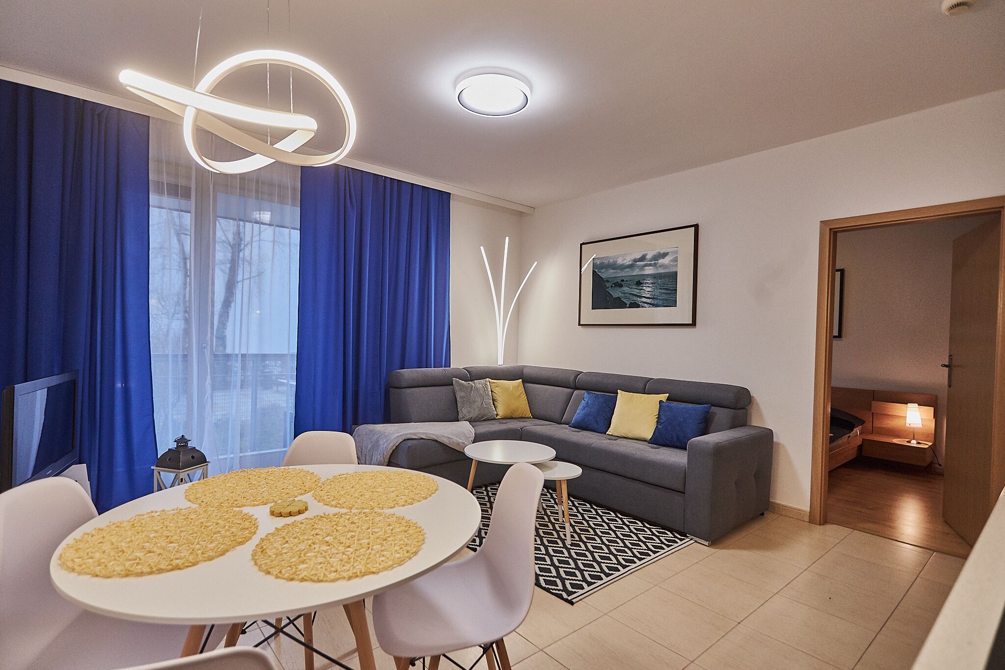 Una sala de estar moderna con un sofá gris, una mesa de comedor redonda con sillas blancas y un gran ventanal con cortinas azules.