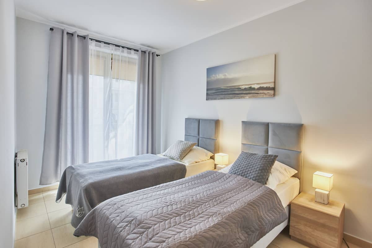 A hotel room with two beds, a painting on the wall, and a window with curtains.