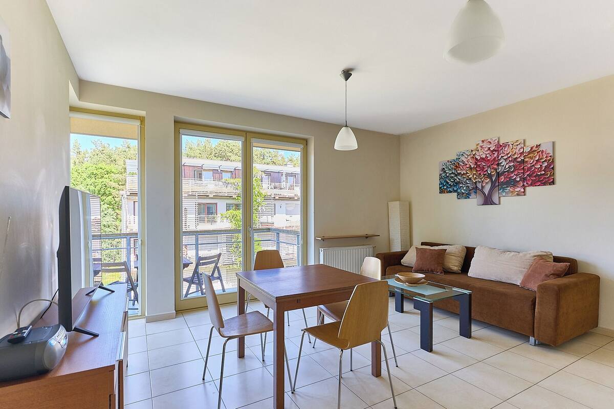 A living room with a dining table, chairs, a sofa, a TV, and a large window with a view of buildings and trees.
