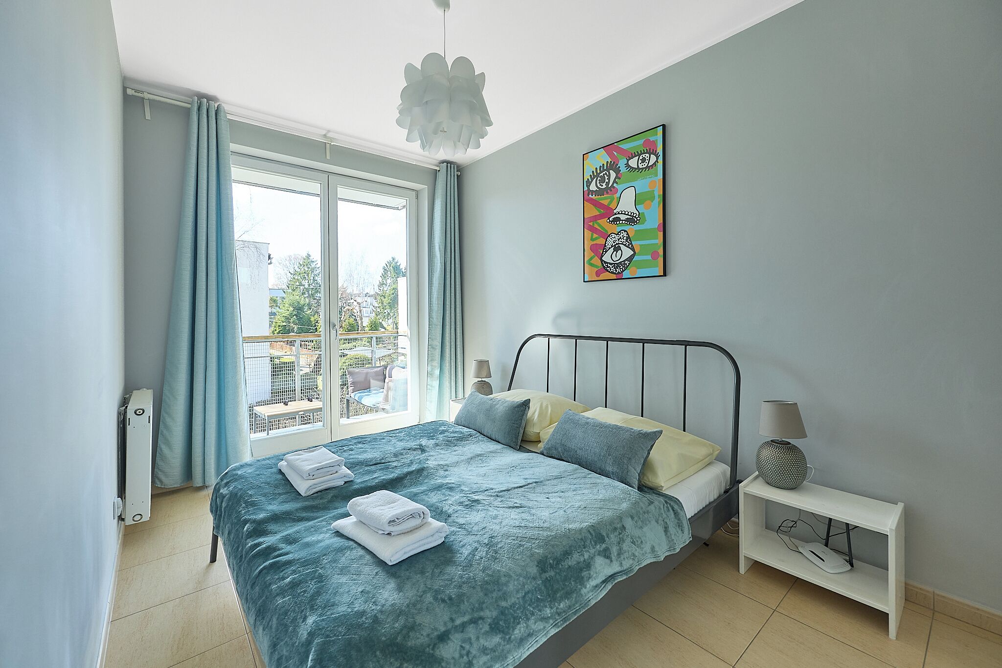 Un dormitorio con cama, una mesita de noche con lámpara, un radiador y una ventana con cortinas.
