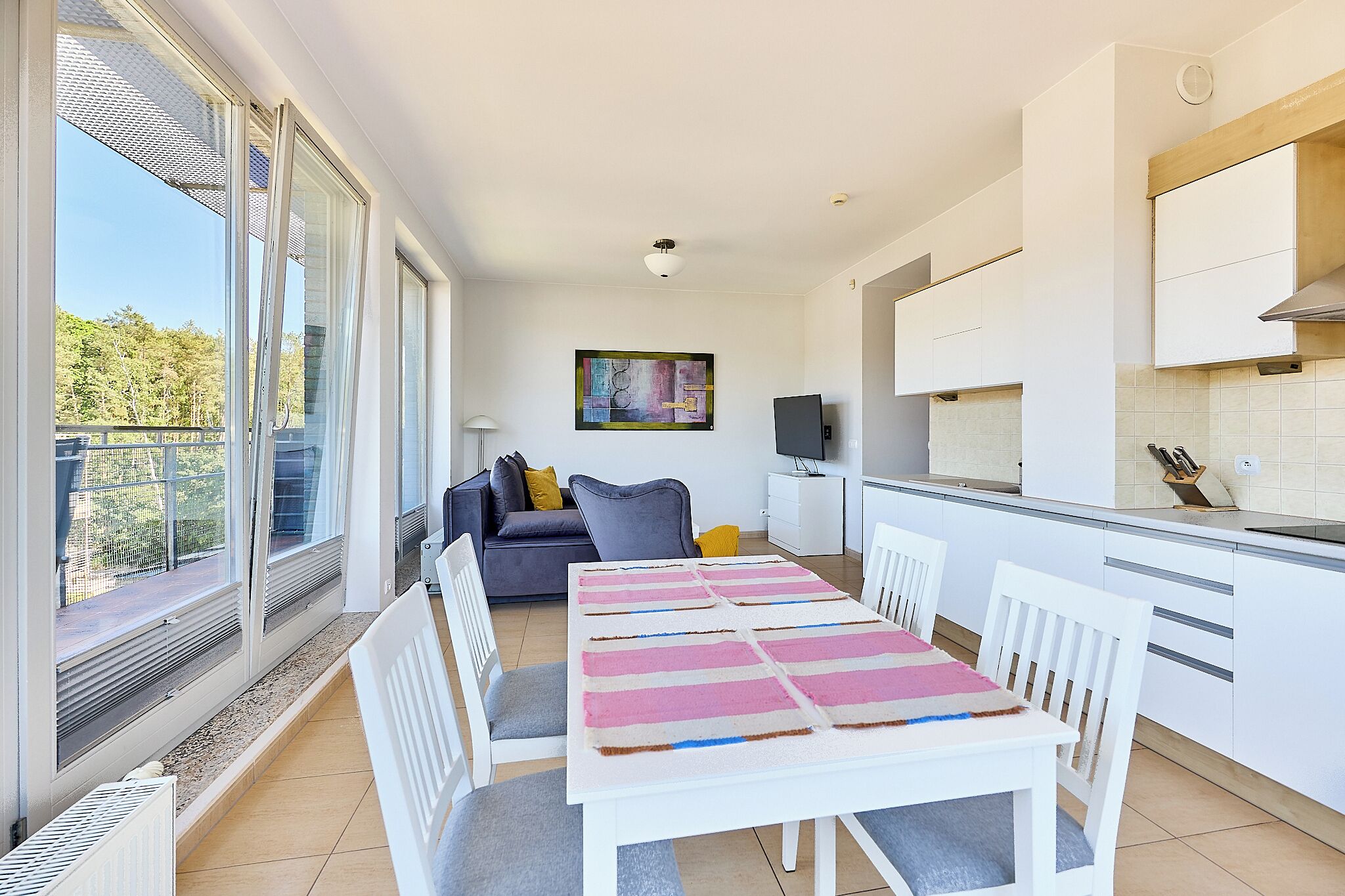 A modern kitchen with white cabinets, a dining table with chairs, a sofa with cushions, a TV, and a balcony with large windows.