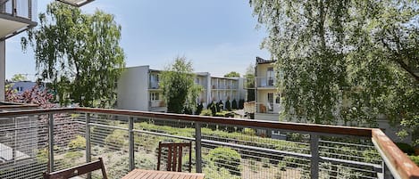Un balcón con mesa y sillas de madera, con vista a un jardín con árboles y una fila de edificios al fondo.