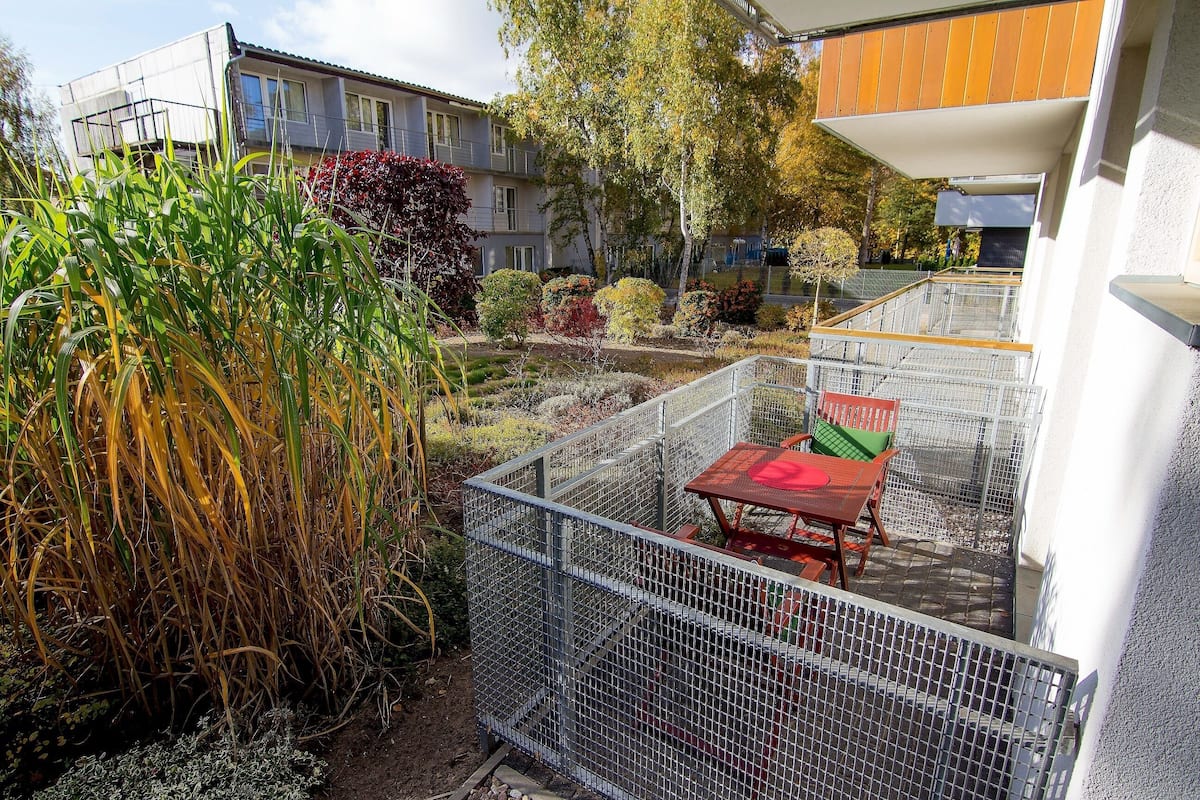A fenced outdoor area with a table and chairs, surrounded by vegetation and a building in the background.