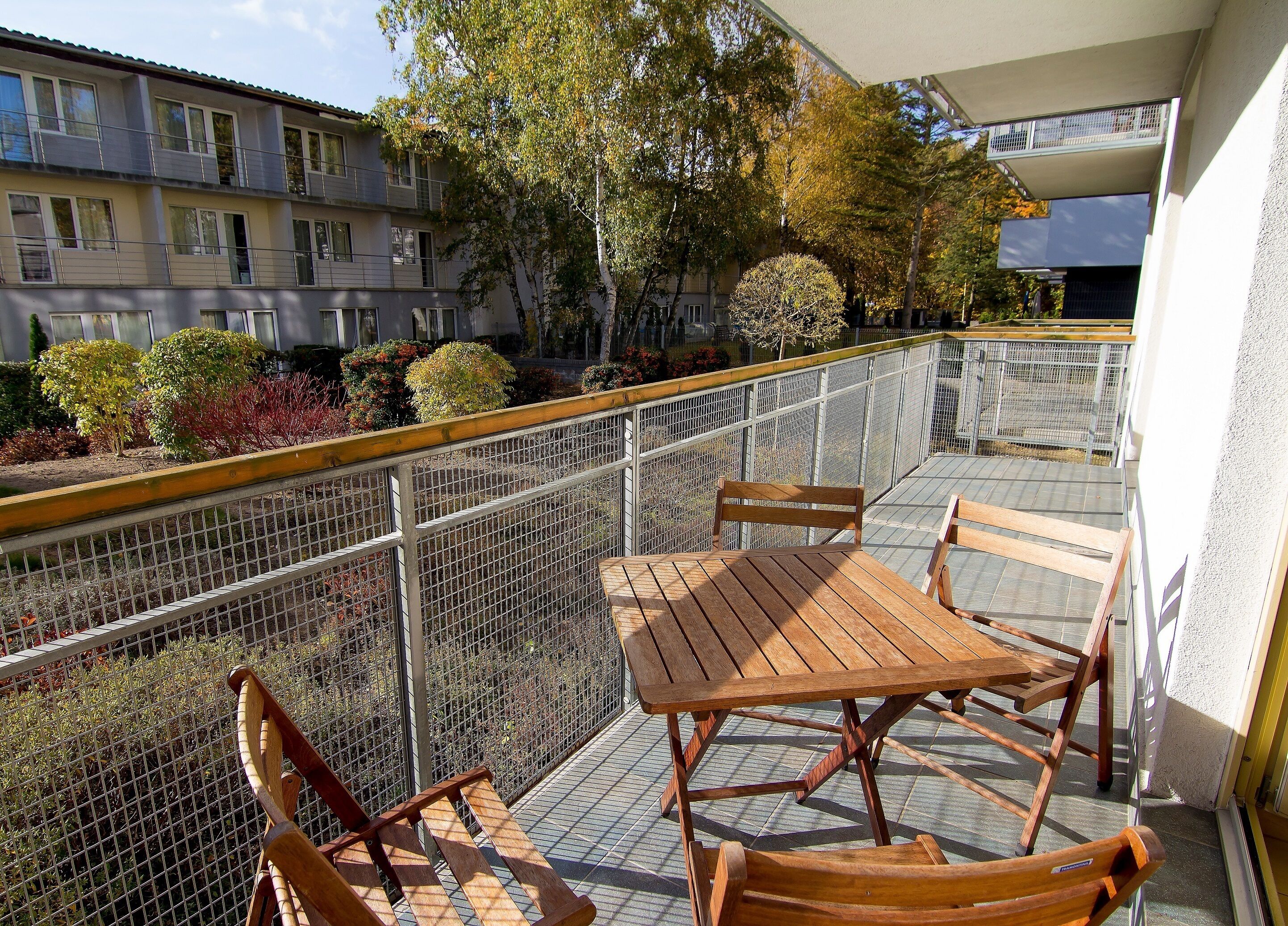 A balcony with a wooden table and chairs, a metal railing, and a view of a building with multiple windows and trees.