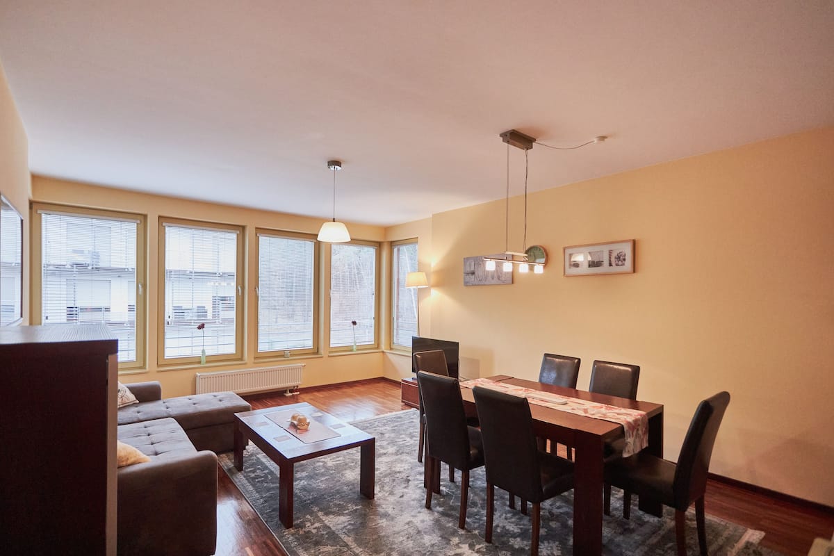 A living room with a sofa, coffee table, dining table with chairs, large windows, and a hanging light fixture.