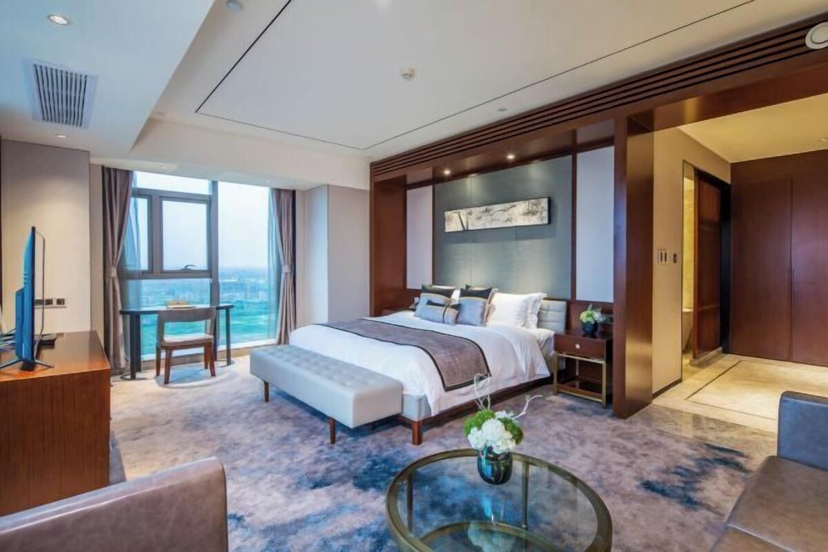 Una habitación de hotel moderna con una cama grande, un escritorio, una silla, una mesa de centro de vidrio y vistas a la ciudad.