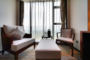 Habitación de hotel con sofá, una silla, una mesita con una botella y un vaso, y un ventanal con cortinas.