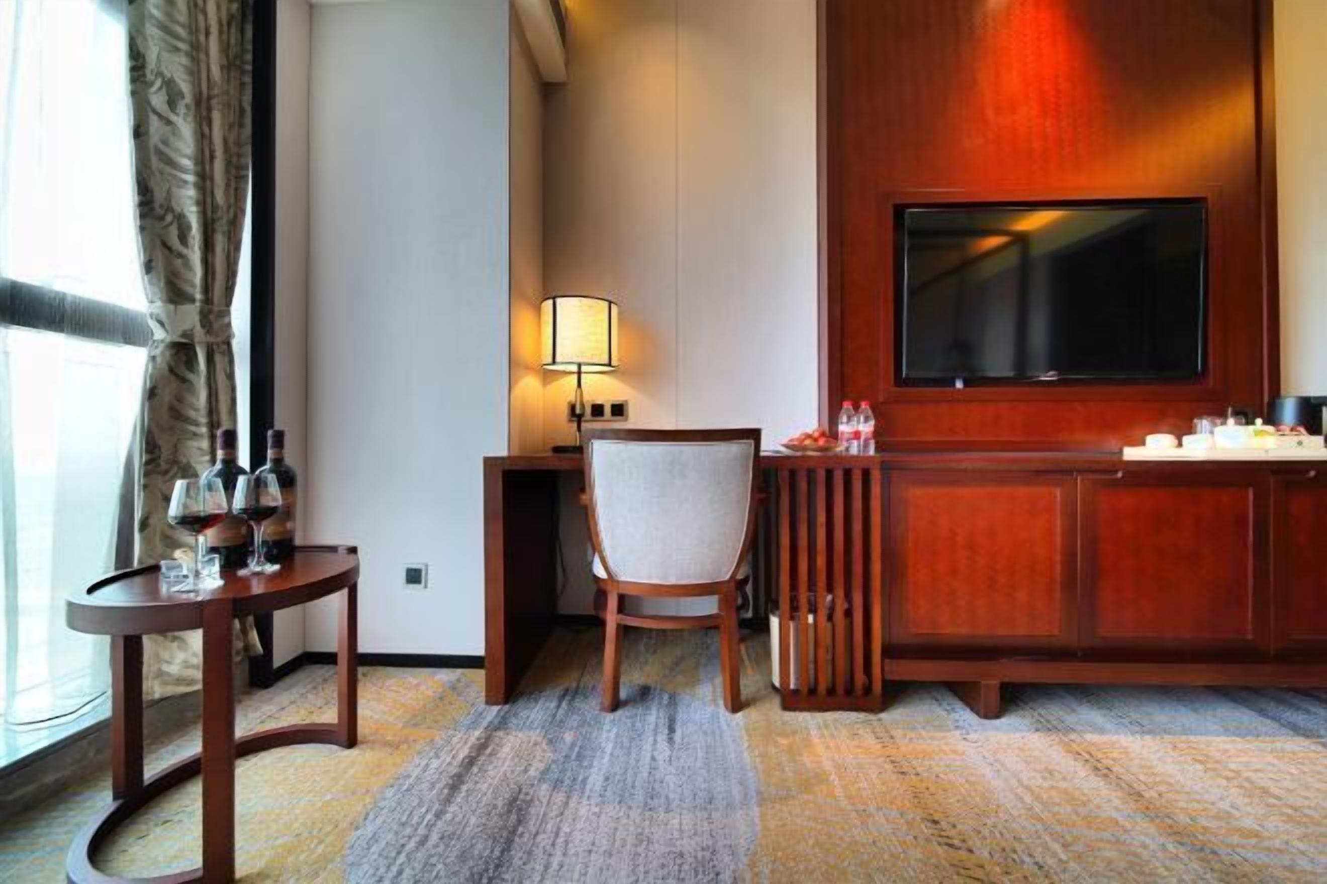 A hotel room with a desk, chair, TV, and a small table with wine bottles and glasses.