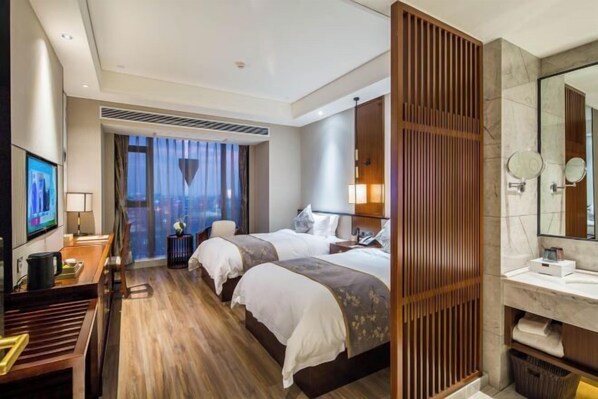 Habitación de hotel con dos camas, un escritorio con televisor, un ventanal grande con cortinas, un baño con espejo y lavabo, y una mampara de madera.