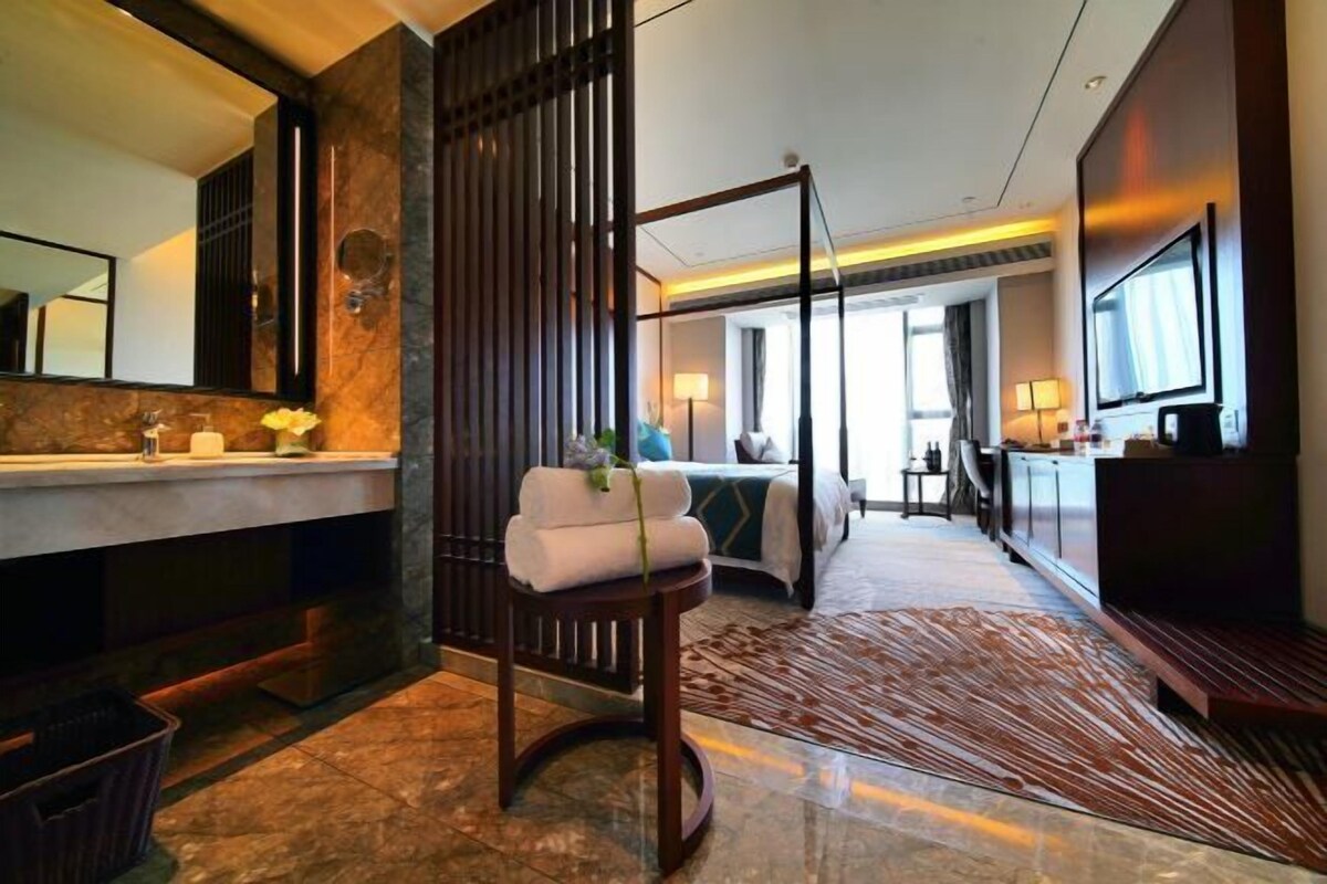 Una habitación de hotel moderna con una cama grande, una zona de estar, un televisor y un baño con un espejo grande y encimera de mármol.