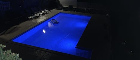 A swimming pool with a blue-lit edge, surrounded by a dark patio area with outdoor furniture.