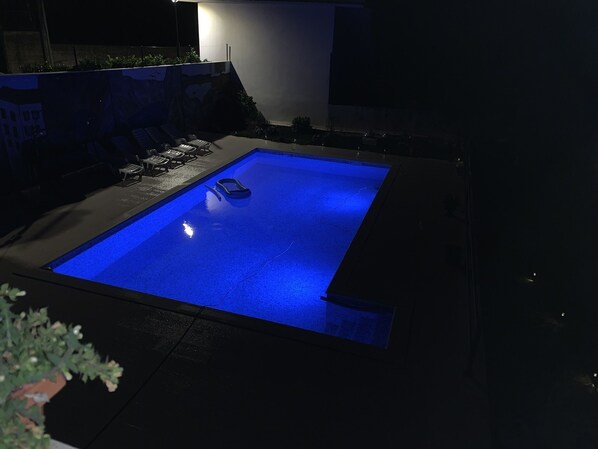 A swimming pool with a blue-lit edge, surrounded by a dark patio area with outdoor furniture.