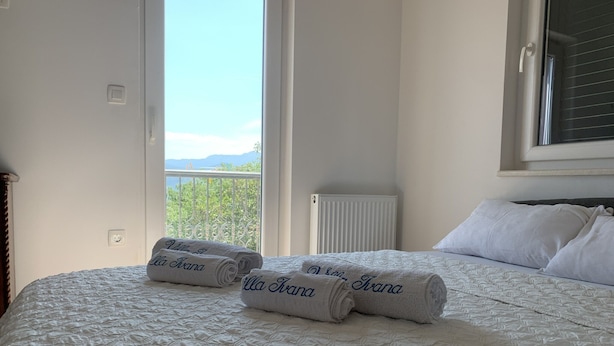 A neatly made bed with a quilted white duvet, two rolled towels with "Villa Ivana" embroidered on them, and a view of a balcony with a railing and greenery. - "imotski Blaue See Apartments , Pool" (Imotski)