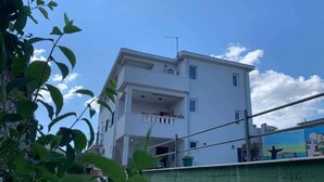 Un edificio residencial de varios pisos con un balcón en el segundo piso, un camino de piedra que conduce a la entrada y una pared verde al frente.