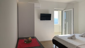 Una habitación de hotel moderna con cama, una pequeña mesa con mantel rojo, televisor montado en la pared y un balcón con vistas.