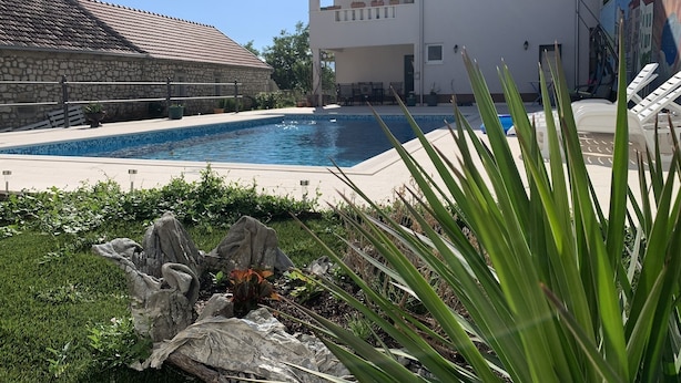 Una piscina rodeada por un muro de piedra, un edificio blanco con balcones y un mural en la pared contigua.