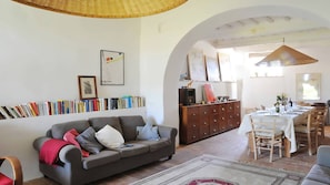 A living room with a sofa, bookshelves, a dining area with a table set for two, and a kitchen visible through an archway. - Casa del Piano Valle dei Calanchi in Front of Civita Di Bagnoregio (Bagnoregio)