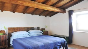 A bedroom with a wooden ceiling, a bed with blue bedding, a nightstand with a lamp, and a window with curtains.