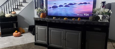 A living room with a TV on a black entertainment center, a staircase, and decorative pumpkins.
