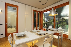 A dining area with a white table set for four, wooden flooring, and large windows offering a view of greenery.