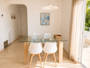 A dining area with a glass table and white chairs, a ceiling light, and a door leading to another room.