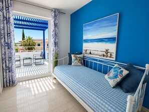A bedroom with a checkered bedspread, a painting of a beach scene, and a view of a balcony with outdoor furniture.