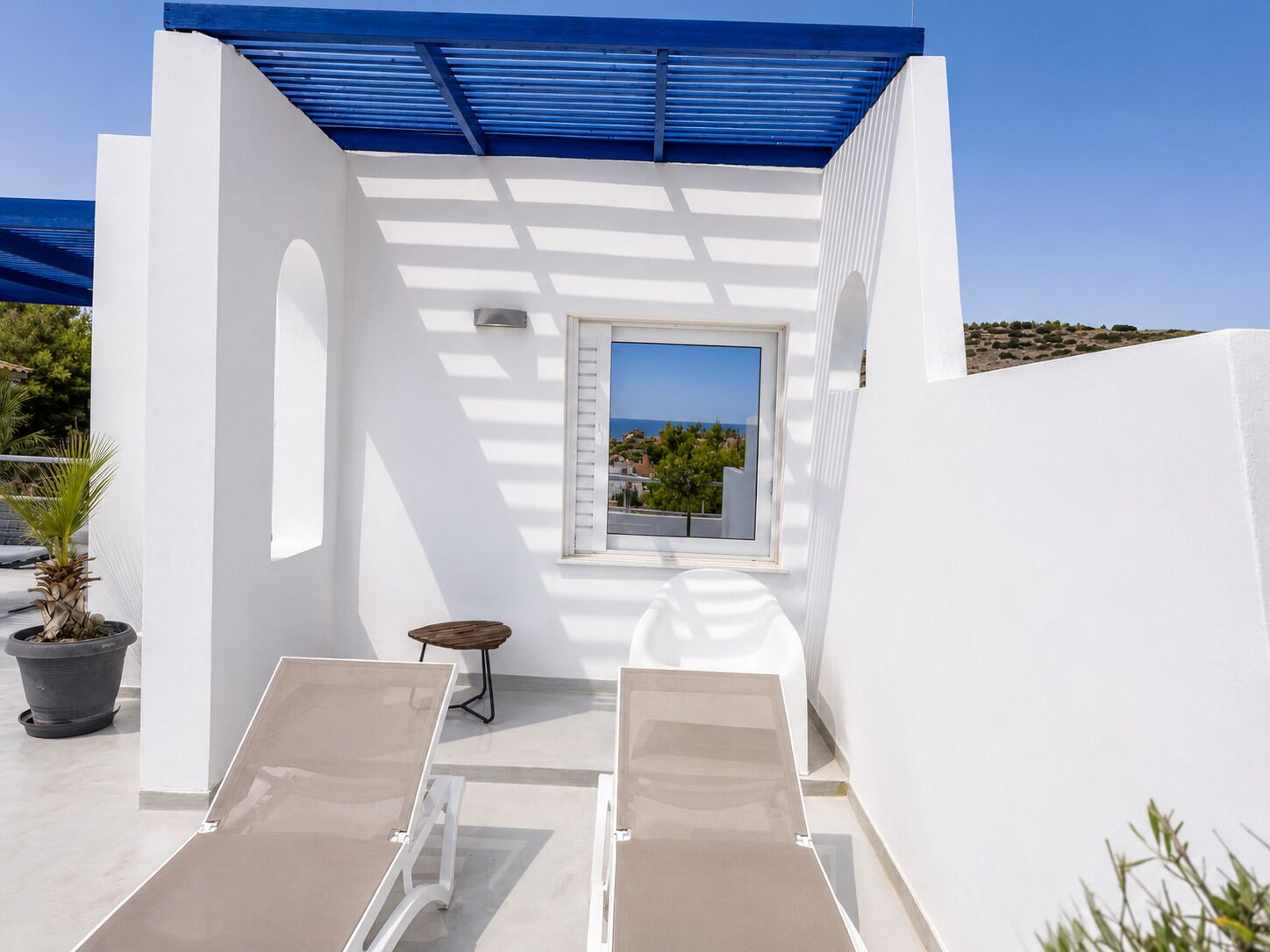 A sunlit outdoor area with two lounge chairs, a small table, and a window overlooking a coastal view.