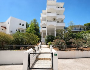 A multi-story white building with a gated entrance, surrounded by greenery and other buildings.
