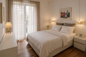 A bedroom with a bed, bedside lamps, a nightstand, a window with sheer curtains, and a framed picture on the wall.