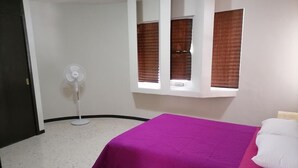 A bedroom with a bed, a window with blinds, a standing fan, and a door.
