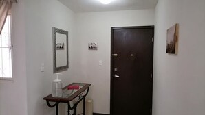 A hallway with a dark door, a small table with a bottle, a mirror, and a picture on the wall.