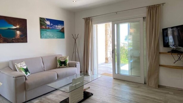 Ein Wohnzimmer mit einer Couch, einem Glastisch und einem Fernseher auf einem Schrank. An der Wand hängen zwei Gemälde und eine Glasschiebetür führt nach draußen.