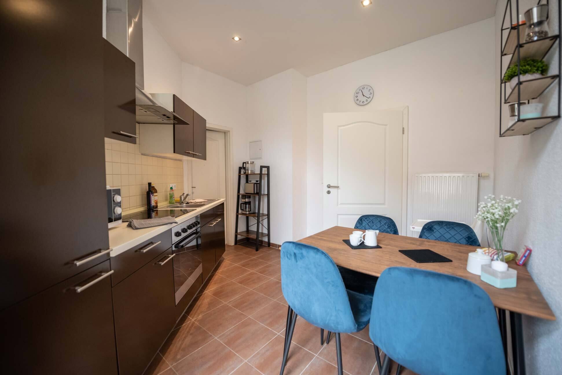 Een moderne keuken met donkere kastjes, een fornuis en een spoelbak. Er is een eethoek met een houten tafel en blauwe stoelen. Op de muur hangt een klok, en een radiator is te zien.
