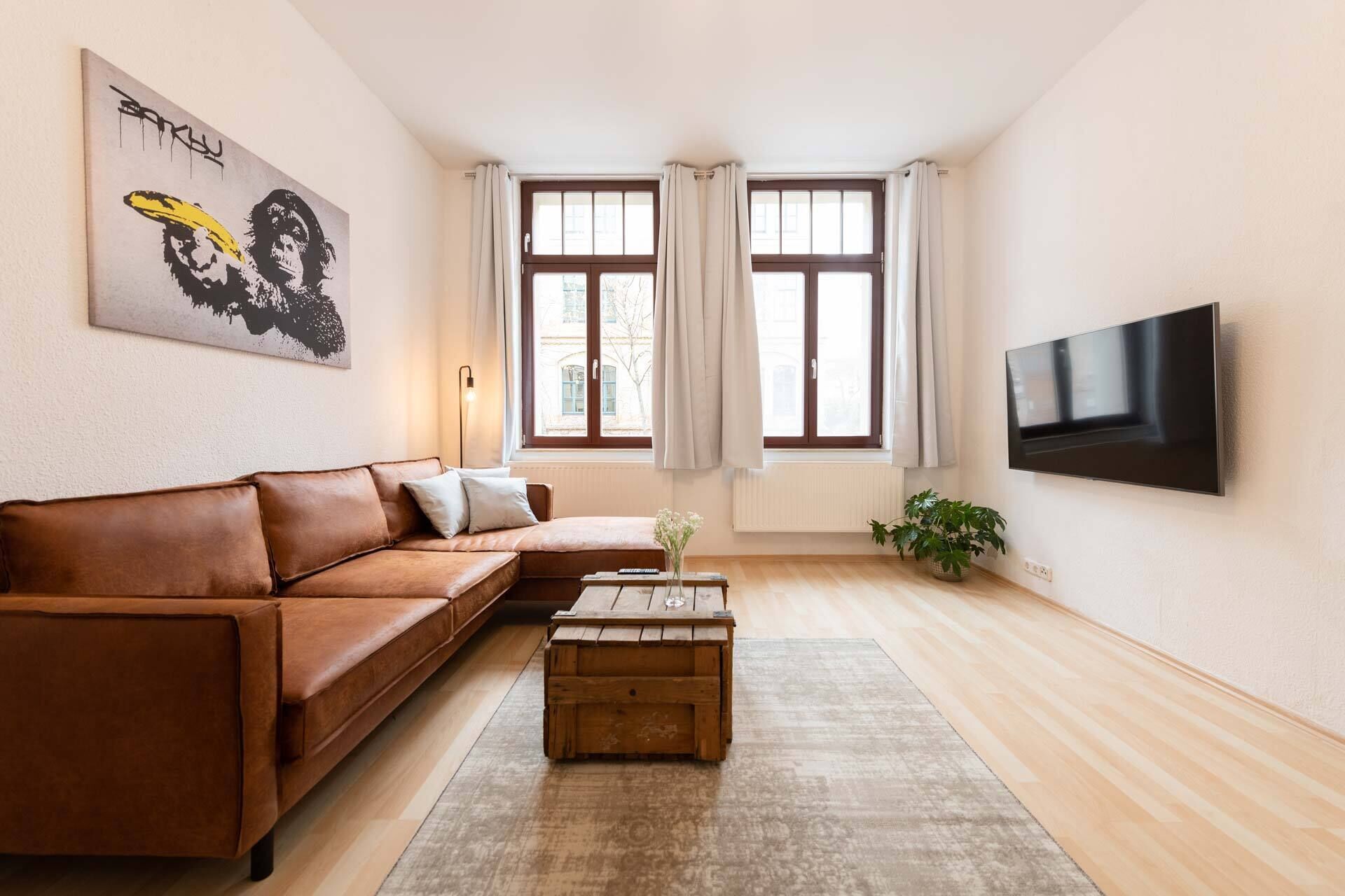 Una sala de estar con un sofá de cuero marrón, una mesa de centro de madera, un televisor montado en la pared y una ventana con cortinas.